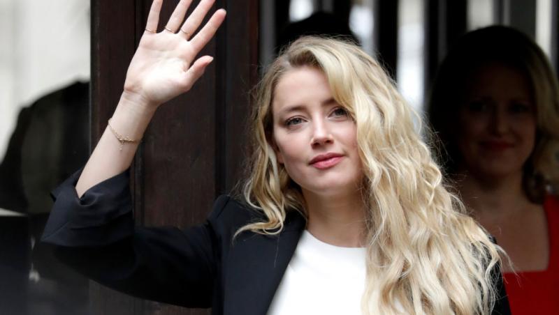 Amber Heard, îmbrăcată într-un tricou alb, peste care poartă un sacou negru, în timp ce salută