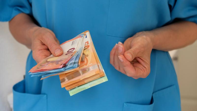 Prim-plan al mâinilor unei asistente într-o uniformă turcoaz care ține și numără bani.