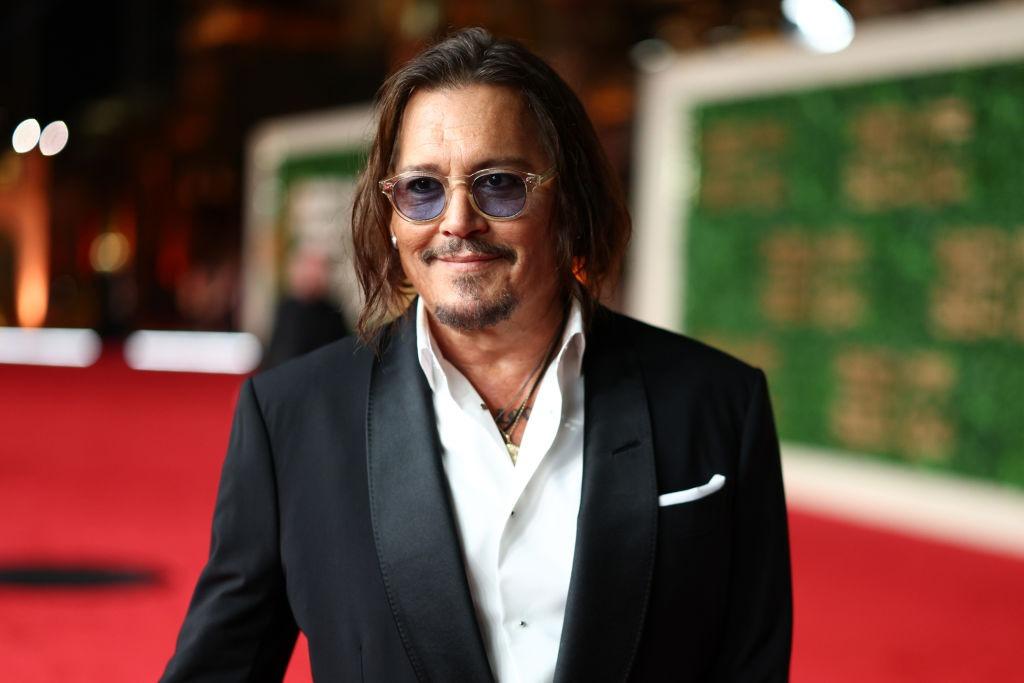 Johnny Depp îmbrăcat cu un costum negru elegant și peste o cămașă albă, accesorizat cu ochelari de soare