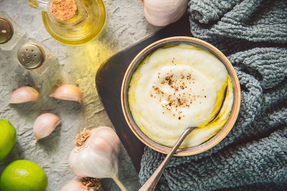 sos toum înt-un bol cu linguriță, cu căței de usturoi, limetă și ulei alături