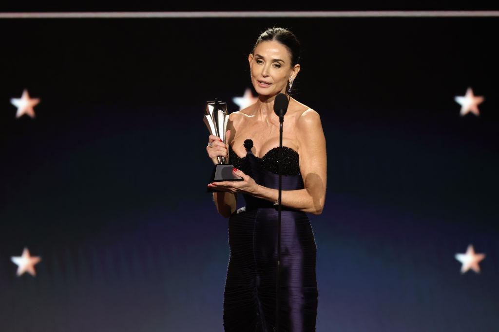 Demi Moore îmbrăcată într-o rochie neagră mulată, ținând un trofeu în mână și vorbind la microfon