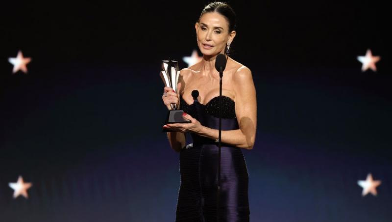 Demi Moore îmbrăcată într-o rochie neagră mulată, ținând un trofeu în mână și vorbind la microfon