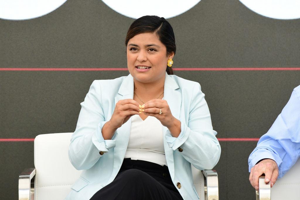 Laura Zapata îmbrăcată cu pantaloni negri, o bluză albă și un sacou cyan