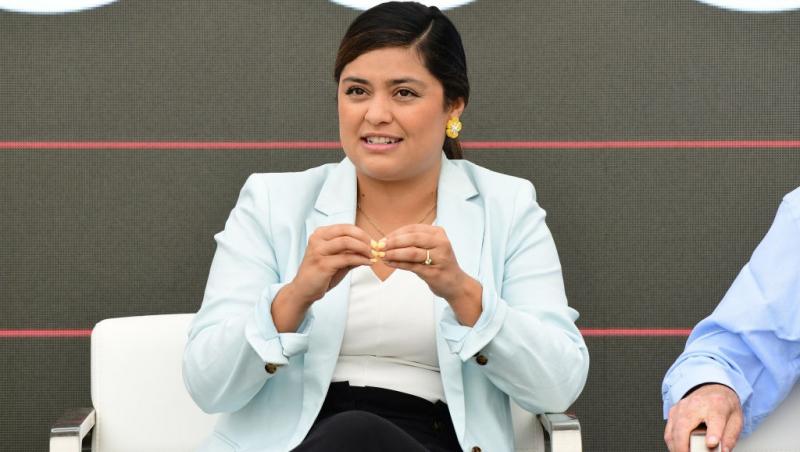 Laura Zapata îmbrăcată cu pantaloni negri, o bluză albă și un sacou cyan