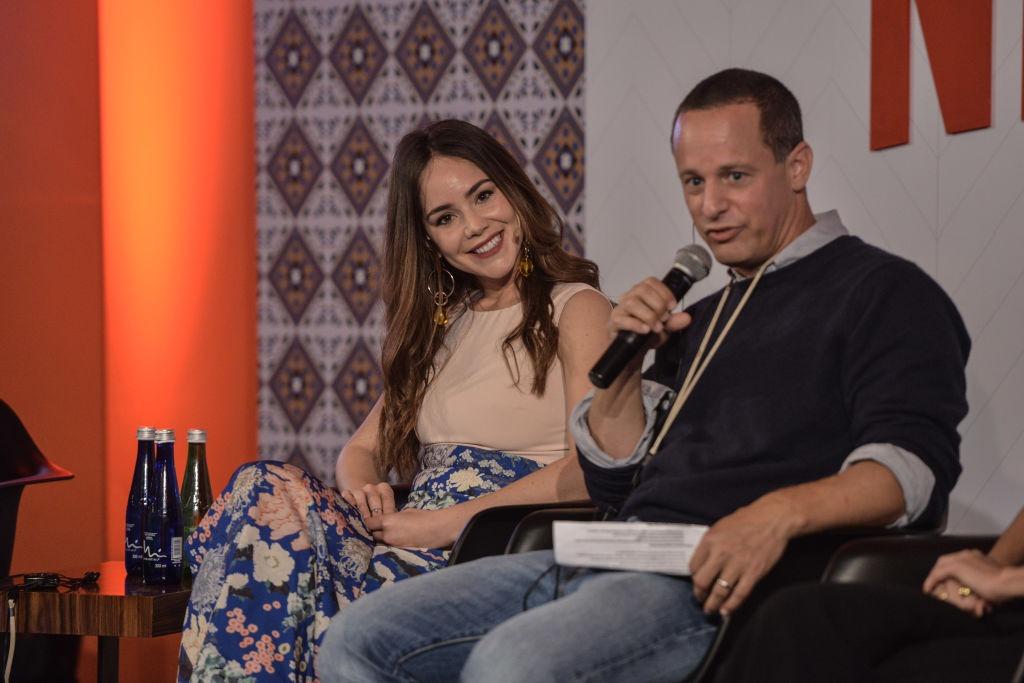 Camila Sodi îmbrăcată cu o fustă florală și un top bej, stând lângă un bărbat care vorbește la microfon