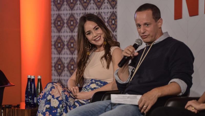 Camila Sodi îmbrăcată cu o fustă florală și un top bej, stând lângă un bărbat care vorbește la microfon