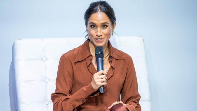 Meghan Markle îmbrăcată într-o cămașă cărămizie, vorbind la microfon