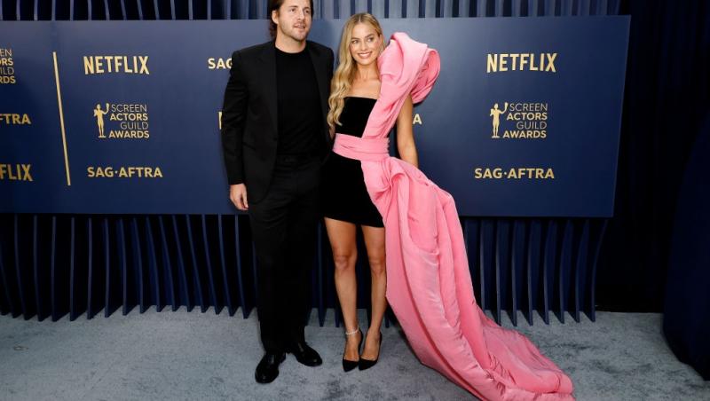 Margot Robbie îmbrăcată cu o rochie neagră cu o fundă roz uriașp, alături de soțul ei, îmbrăcat în negru