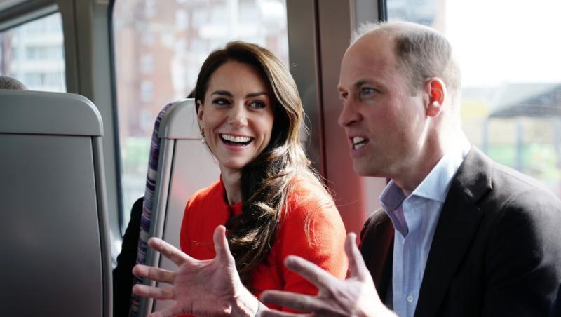 Prințul William îmbrăcat într-un costum negru, alături de Prințesa Kate, îmbrăcată într-o rochie roșie, discutând într-un autobuz