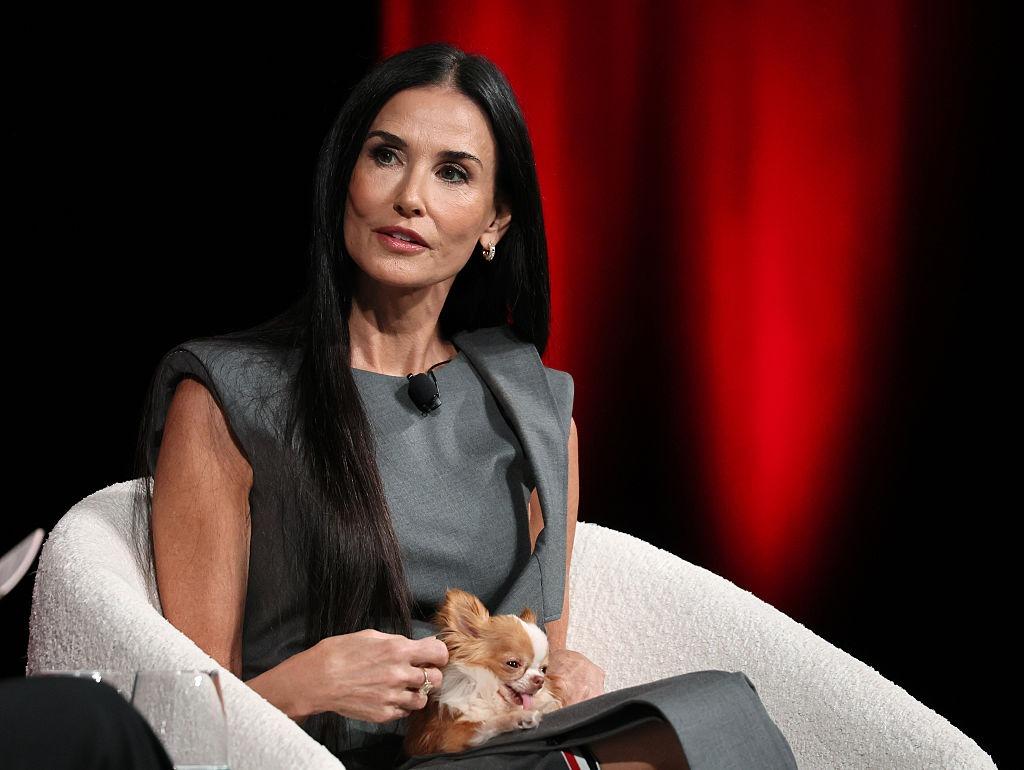 Demi Moore îmbrăcată într-o rochie gri, fără mâneci, stând pe un fotoliu alb cu un cățeluș în brațe