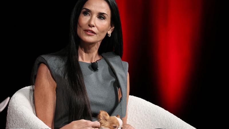 Demi Moore îmbrăcată într-o rochie gri, fără mâneci, stând pe un fotoliu alb cu un cățeluș în brațe
