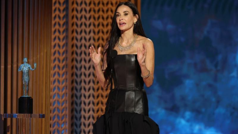 Demi Moore îmbrăcată într-o rochie neagră, vorbind la microfon pe scenă