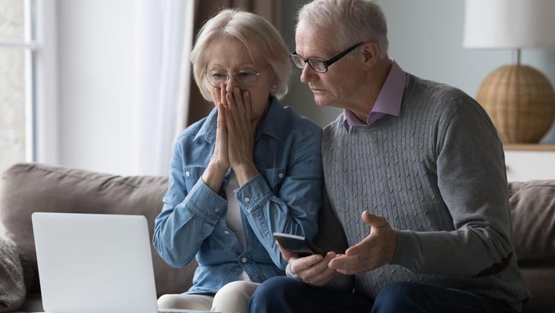 Doi seniori care se uită la laptop și la telefon și încearcă să își dea seama cum au piedut bani