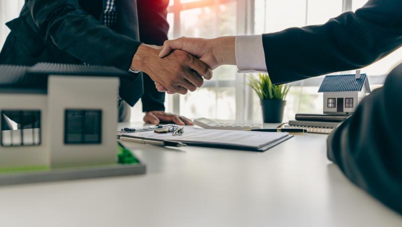 bărbați la costum ce își dau mâna peste niște acte și machete de case