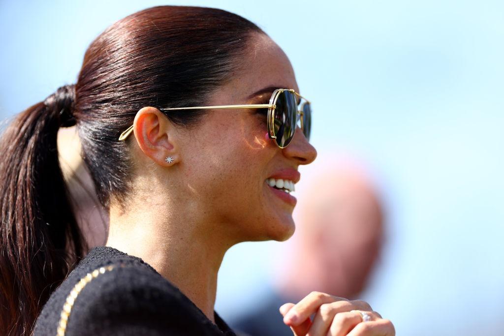 Meghan Markle îmbrăcată într-o rochie neagră elegantă și ochelari de soare, zâmbind fericită