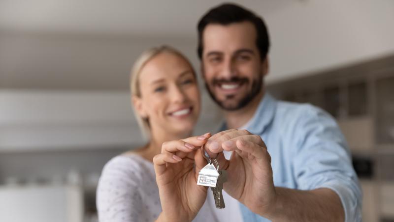 Un bărbat brunet cu barbă și o femeie blondă care țin &icirc;n m&acirc;ini cheile de la imobil și un breloc cu o casă