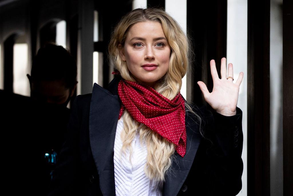 Amber Heard îmbrăcată cu un sacou negru accesorizat cu o eșarfă roșie cu buline albe, ținând mâna stângă ridicată