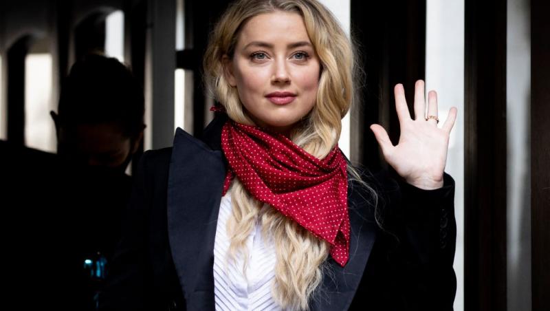 Amber Heard îmbrăcată cu un sacou negru accesorizat cu o eșarfă roșie cu buline albe, ținând mâna stângă ridicată