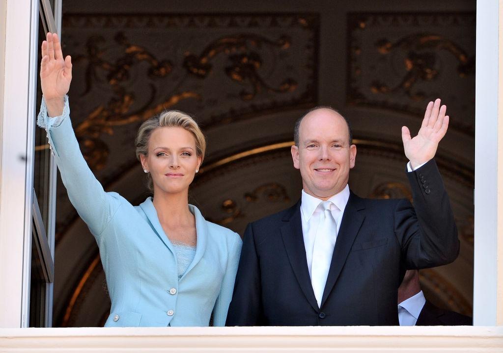 Prințul Albert și ai Prințesa Charlene, ambii îmbrăcați elegant, stând la fereastră și salutând oamenii