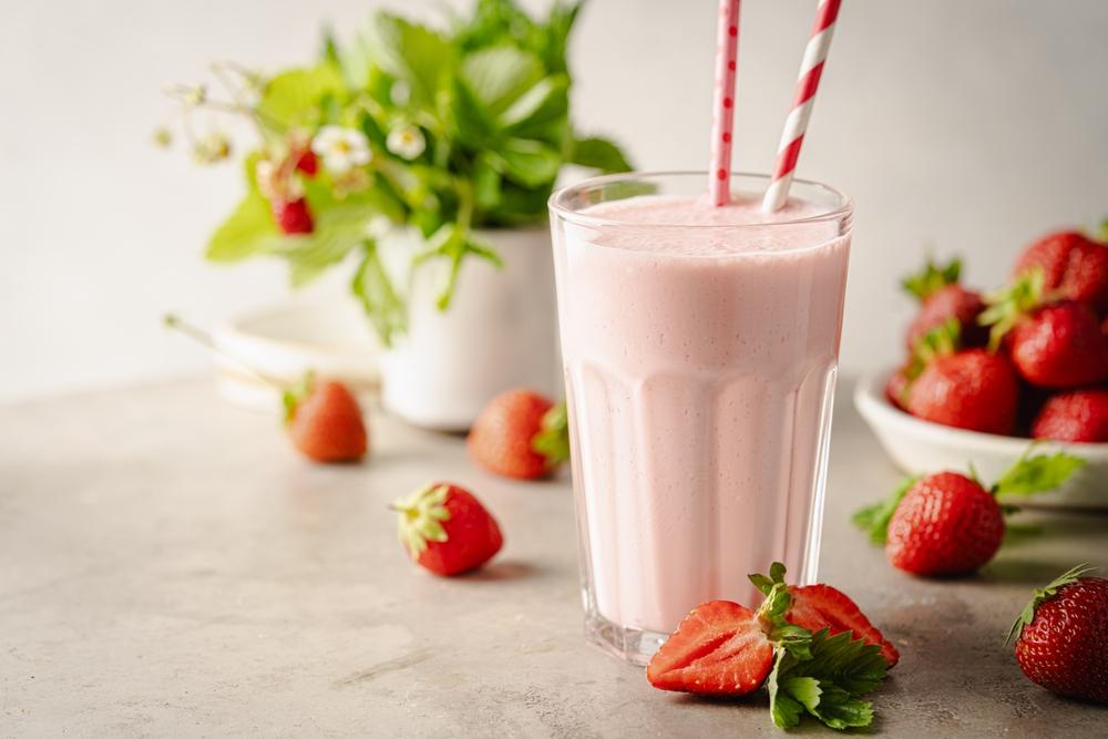 pahar înalt cu milkshake de căpșuni, cu pai și căpșuni proaspete alături