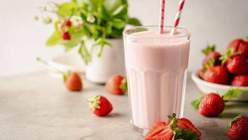 pahar înalt cu milkshake de căpșuni, cu pai și căpșuni proaspete alături