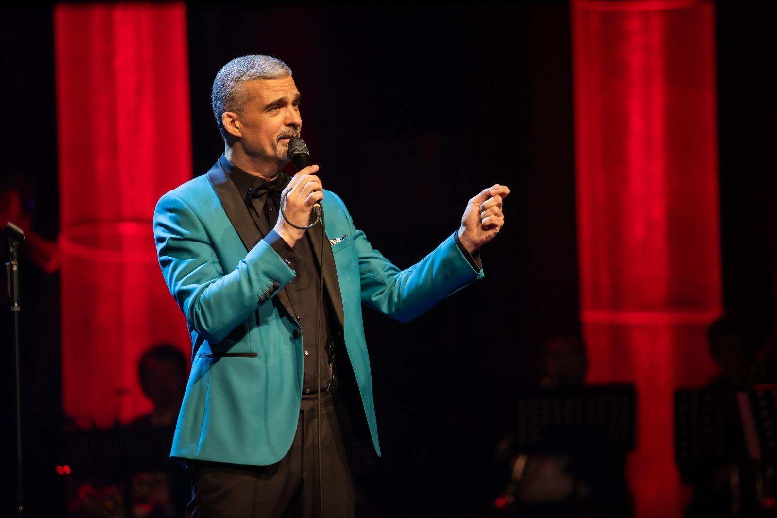 Aurelian Temișan îmbrăcat cu un costum bleu cu revere negre, cântând la microfon