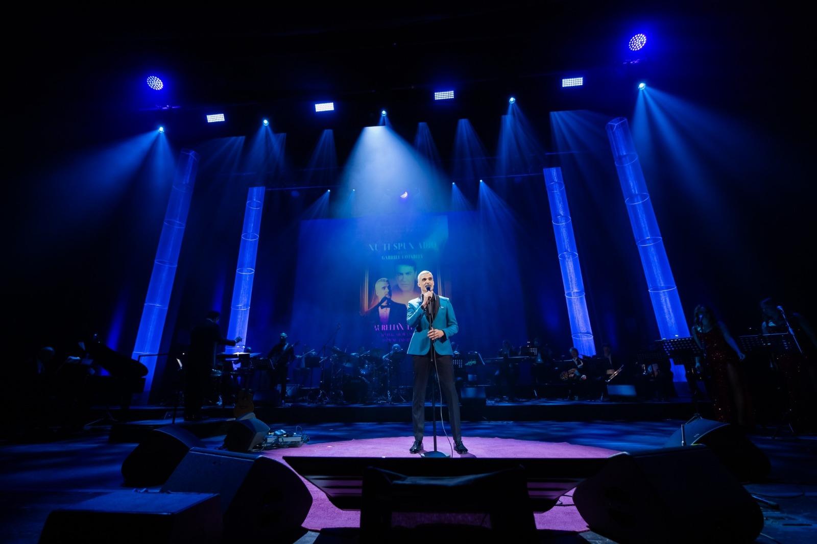 Aurelian Temișan îmbrăcat cu un costum bleu cu revere negre, cântând la microfon