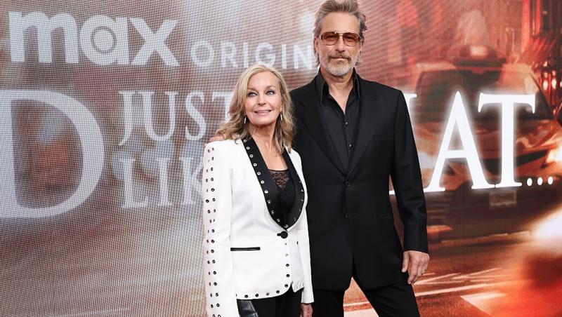 Bo Derek îmbrăcată cu un sacou alb, alături de soțul ei, îmbrăcat într-un costum negru