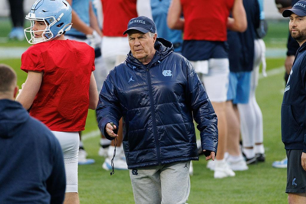 Bill Belichick îmbrăcat cu o geacă lucioasă, pe terenul echipei New England Patriots