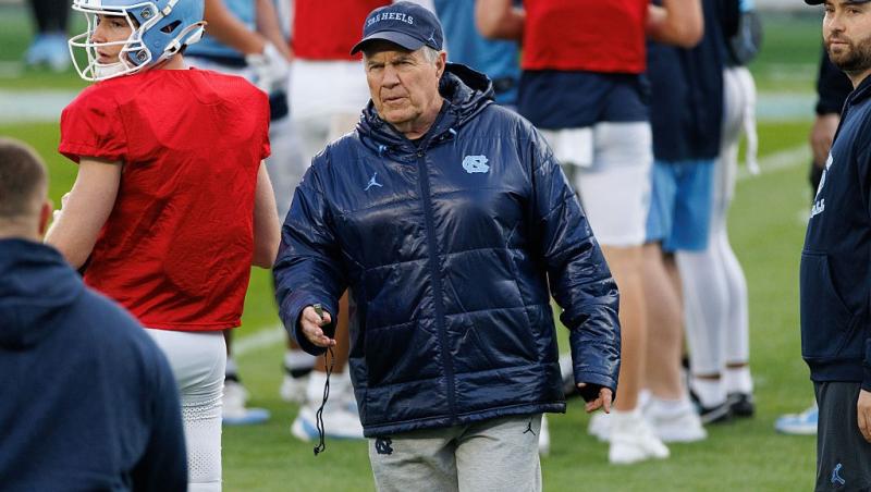 Bill Belichick îmbrăcat cu o geacă lucioasă, pe terenul echipei New England Patriots
