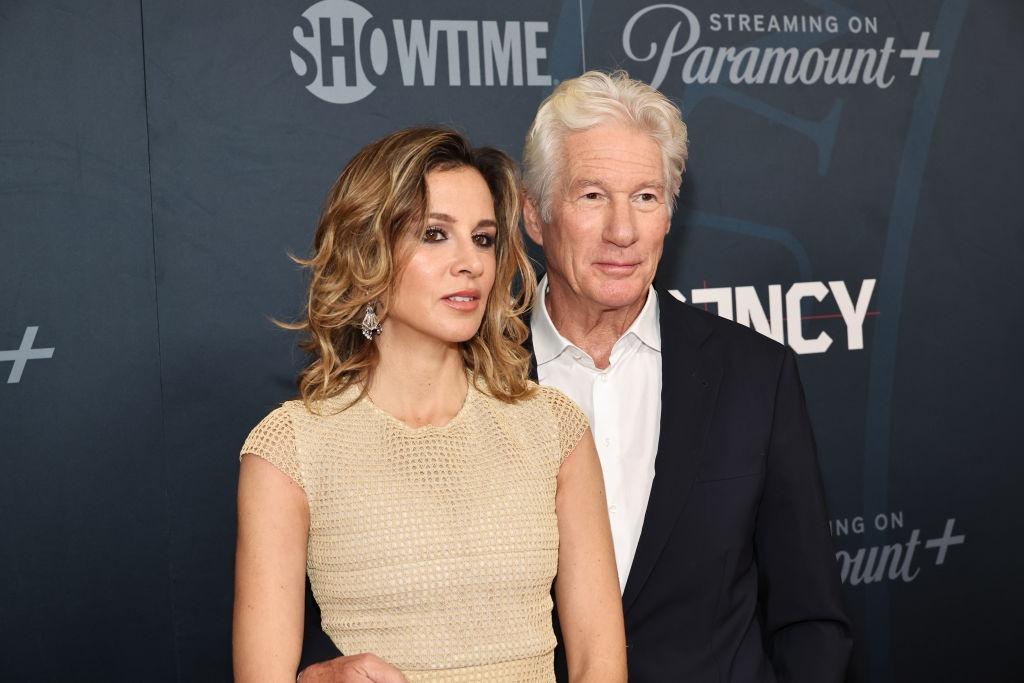 Richard Gere îmbrăcat cu un costum negru peste o cămașă albă, alături de soția lui, îmbrăcată cu o rochie bej