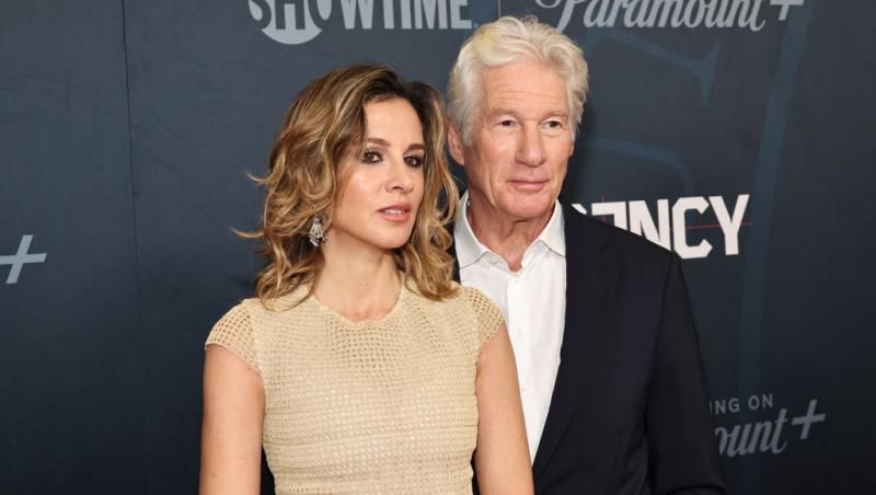 Richard Gere îmbrăcat cu un costum negru peste o cămașă albă, alături de soția lui, îmbrăcată cu o rochie bej