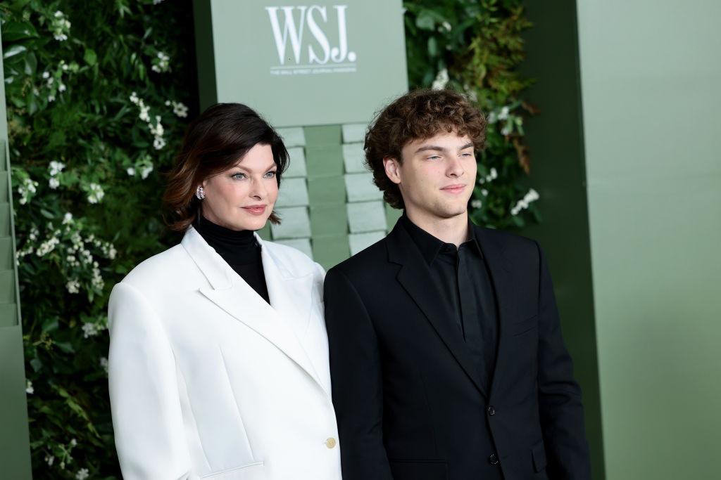 Linda Evangelista îmbrăcată cu un sacou alb peste o bluză neagră, alături de fiul ei, îmbrăcat în negru