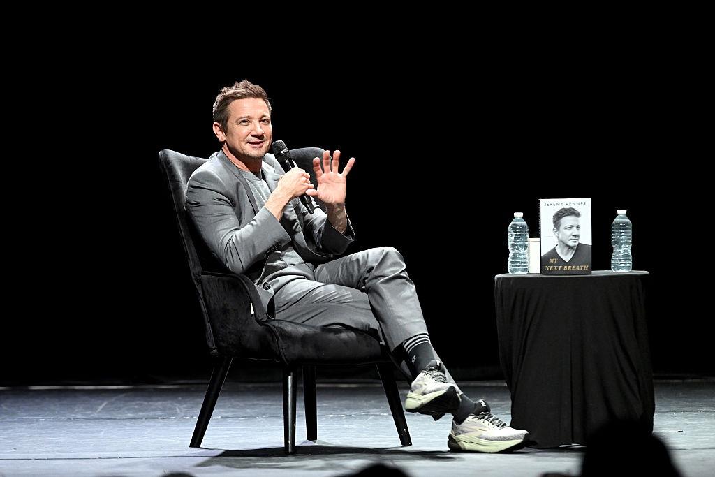 Jeremy Renner îmbrăcat într-o ținută gri, vorbind la microfon