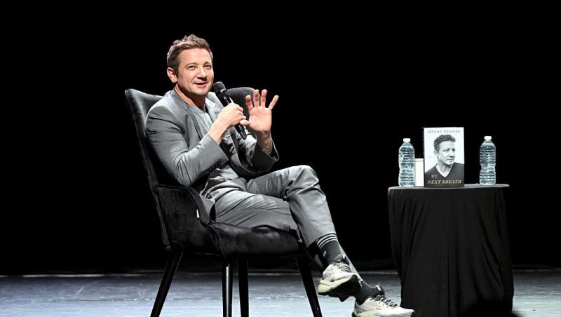 Jeremy Renner îmbrăcat într-o ținută gri, vorbind la microfon