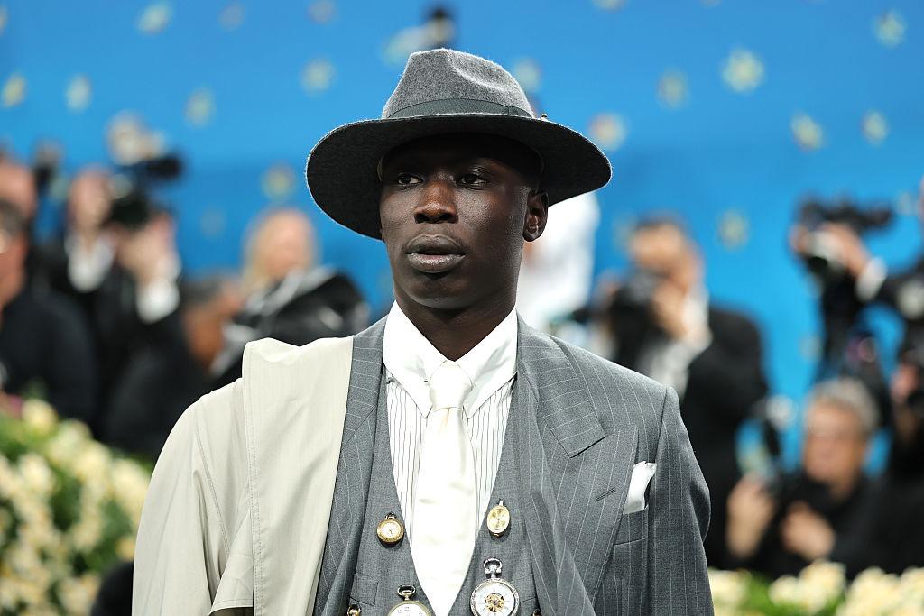 Khaby Lame îmbrăcat cu un costum gri elegant, accesorizat cu o pălărie și mai multe ceasuri