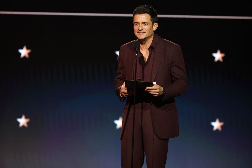 Orlando Bloom îmbrăcat cu un costum elegant, vișiniu, vorbind la microfon pe scenă