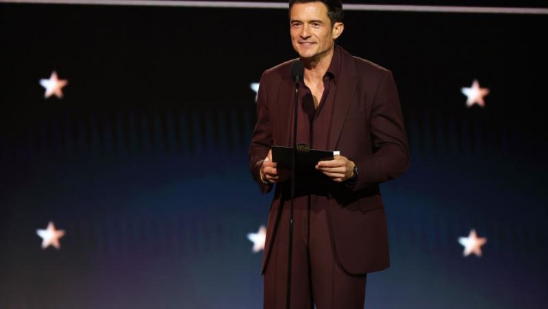 Orlando Bloom îmbrăcat cu un costum elegant, vișiniu, vorbind la microfon pe scenă