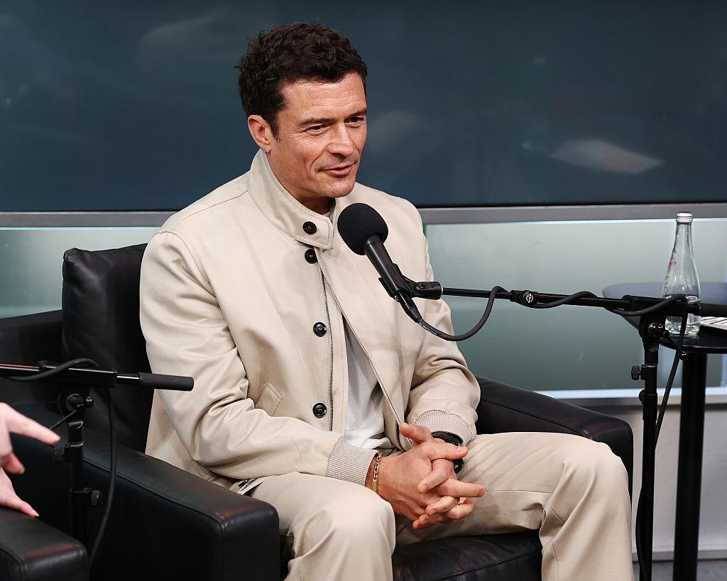 Orlando Bloom îmbrăcat cu o ținută bej, vorbind la microfon