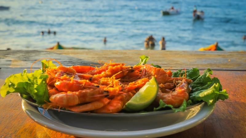 Creveți într-o farfurie cu verdeață și lime, iar pe fundal se vede marea