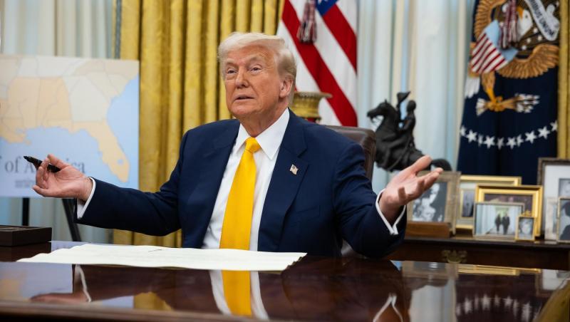 Donald Trump într-un sacou albastru, cămașă albă și cravată galbenă ținând în mână un stilou