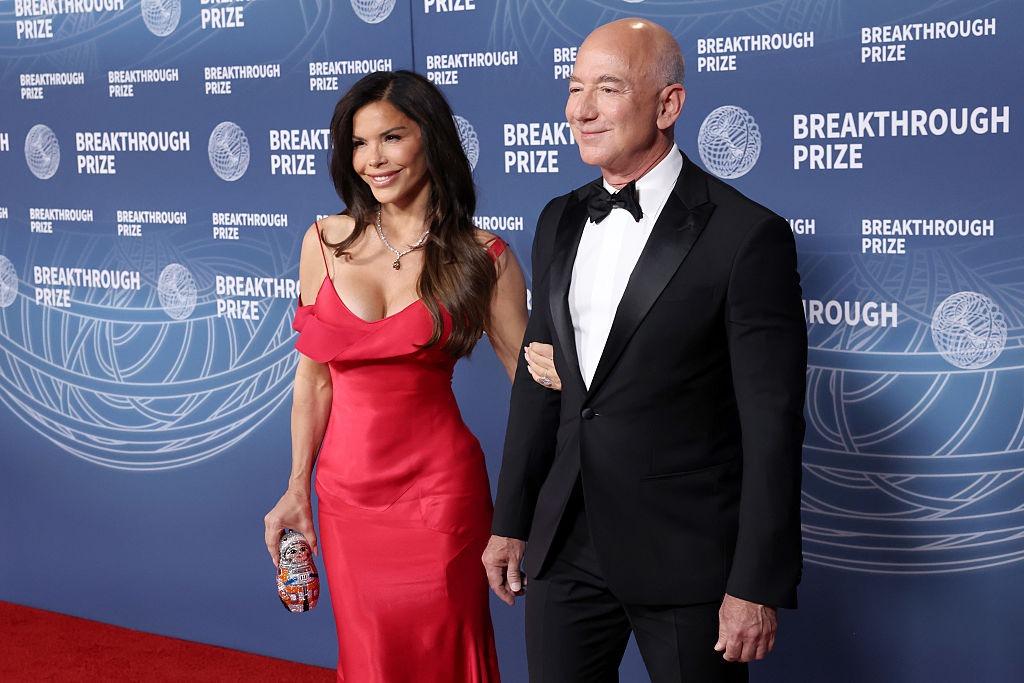Jeff Bezos îmbrăcat într-un costum negru, alături de Lauren Sanchez, îmbrăcată într-o rochie roșie