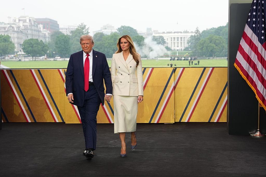 Donald Trump îmbrăcat cu un costum bleumarin și o cravată roșie, mergând alături de soția lui