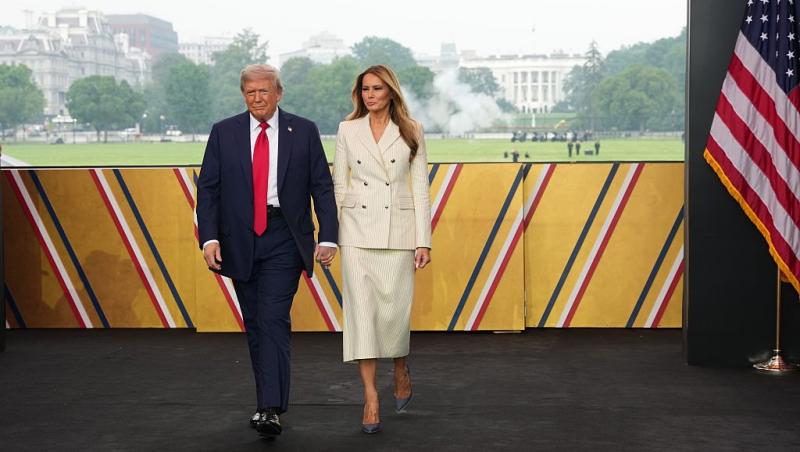 Donald Trump îmbrăcat cu un costum bleumarin și o cravată roșie, mergând alături de soția lui