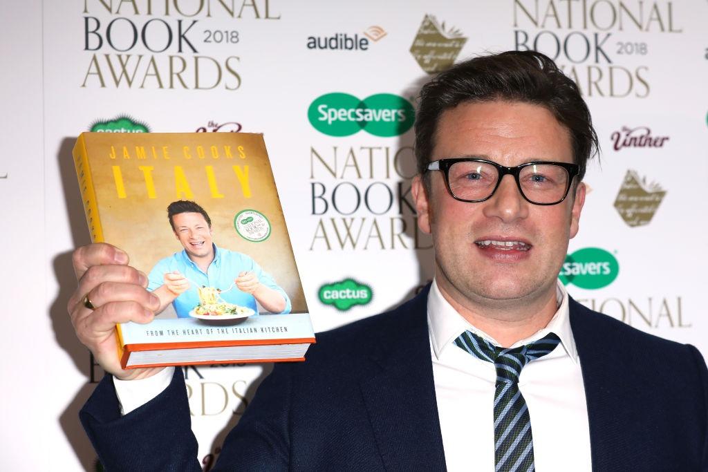 Jamie Oliver îmbrăcat cu un costum negru, ținându-și cartea de bucate în mână