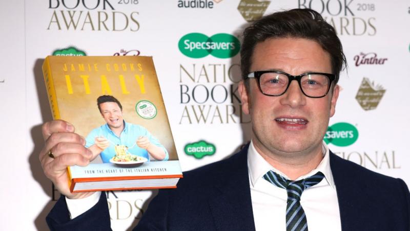 Jamie Oliver îmbrăcat cu un costum negru, ținându-și cartea de bucate în mână