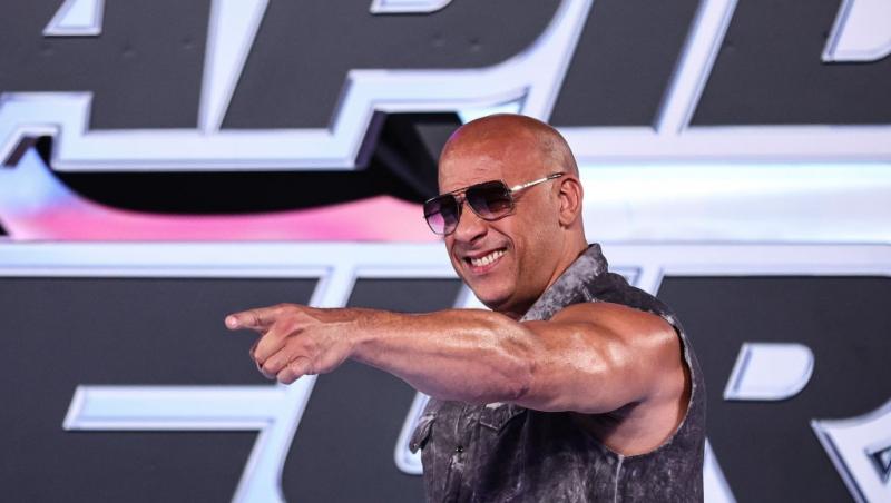 Vin Diesel purtând o jachetă gri și ochelari de soare, zâmbind