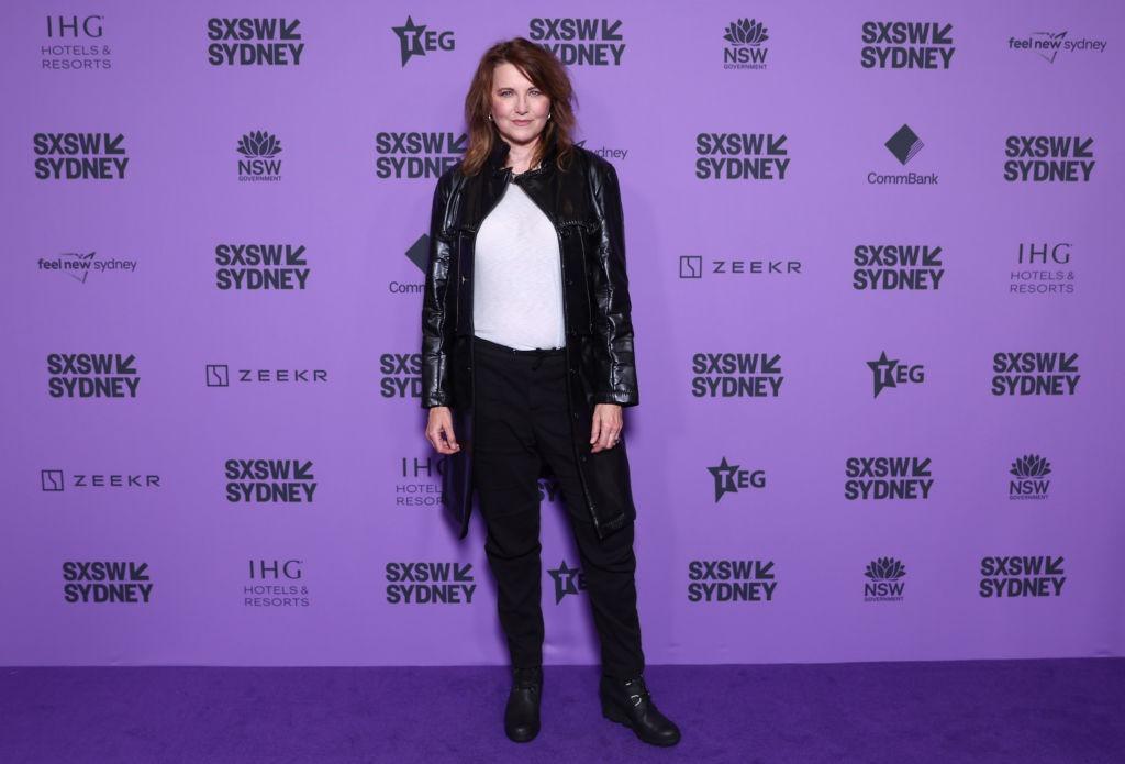 Lucy Lawless îmbrăcată cu o jachetă neagră din piele, peste o bluză albă