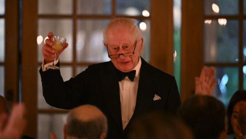 Regele Charles îmbrăcat cu un costum negru elegant, ținând în mână un pahar cu băutură