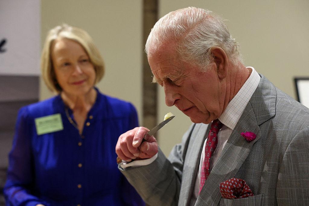 Regele Charles îmbrăcat cu un costum gri, mirosind o bucată de brânză